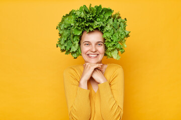 Cute smiling woman wearing casual shirt and lettuce wreath standing isolated over yellow background keeps hands under chin looking at camera with charming smile.