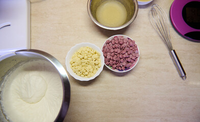 Top view marble kitchen countertop with ingredients for making a mousse cake. Whipped cream in food processor bowl, whisk and chips of white and ruby pink organic confectionery chocolate. Still life
