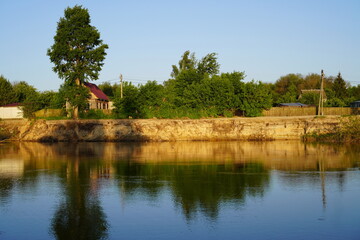 summer landscape with a tall tree near a country house on the bank of a river cliff with reflections against the blue sky