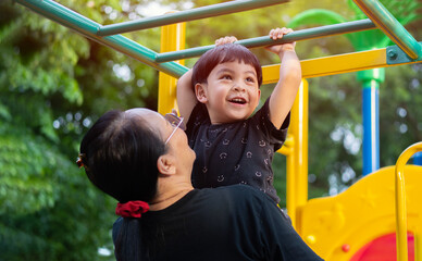 Portrait of a funny little Asian boy hangs by horizontal bar with mother on playground in the sunny...