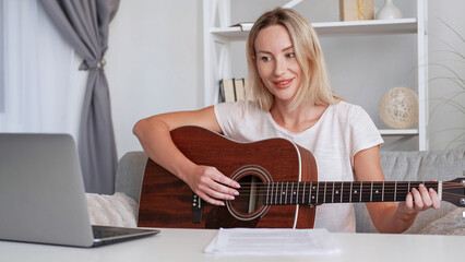 Online guitar class. Music tutor. Smiling pleased woman teacher tutorial lesson teaching playing instrument recording educational video on laptop camera home interior.