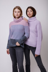 Two fashion models in pastel lilac and pink sweater, blouse, black jeans, denim. Beautiful young woman. Studio shot, portrait. White background. Asian and  blonde. Slim figure