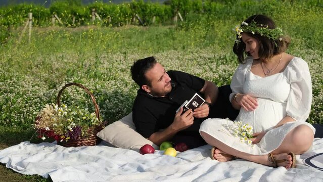 In slow motion, a pregnant woman with a crown of daisies and her partner in black sit on a picnic blanket on the grass, holding their baby's ultrasound images, immersed in serene anticipation