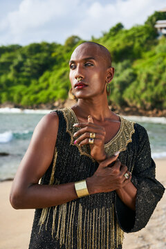 Flamboyant gay black man with luxury golden jewelry poses on scenic ocean beach. Gender fluid ethnic fashion model brass rings, bracelet, earrings, accessories looks stands with sophisticated posture.