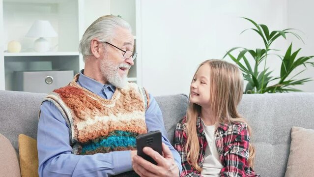 Grey-bearded aged male showing pictures to beloved grandchild on personal wireless gadget. Delightful grandpa and girl enjoying viewing files together and spending great time.
