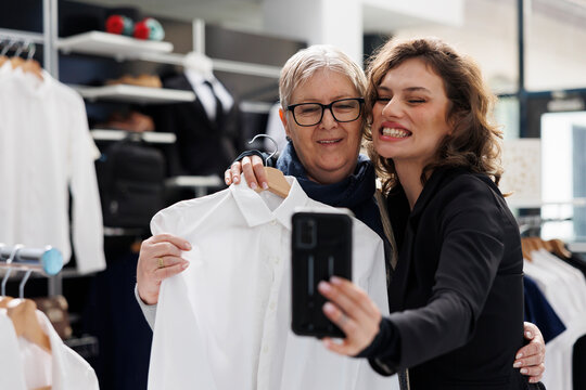 Smiling women taking selfie with mobile phone in shopping centre, elderly woman holding white shirt in front of camera. Shopaholic customers having fun in modern boutique while buying clothes