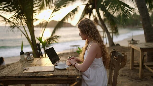 Female cryptocurrency trader at laptop checks charts online working remotely at outdoor tropical seaside cafe at sunset. Woman crypto broker analyses graphic of stock exchange rates by the ocean