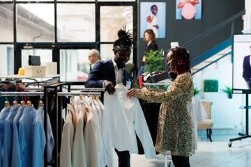 Showroom manager helping client with white shirt, discussing clothes fabric in shopping centre. African american pregnant woman buying fashionable maternity merchandise. Fashion concept