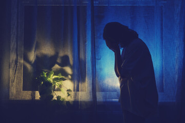 Woman crying at night dark window, low key silhouette. The shadow of a sad female against the background of a window curtain.