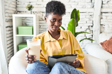 African woman enjoys breakfast and a video on her tablet, sipping milk in her living room.