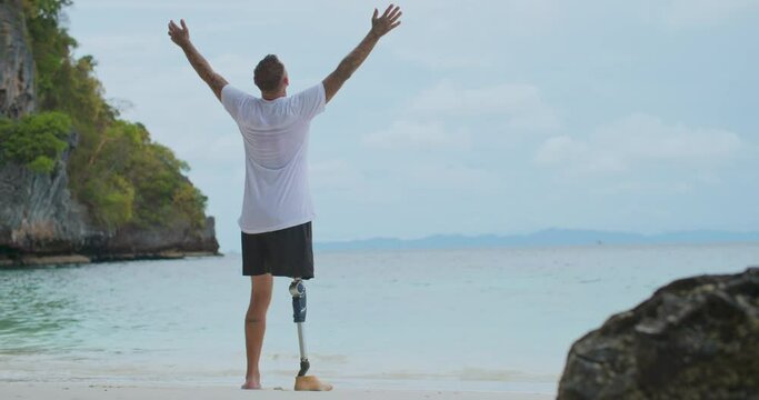 Back view adult male with artificial bionic leg prosthesis walking and raising hands to the sky by the sea. Disability caucasian man lifting arm up enjoying scenic landscape in summer vacation travel.