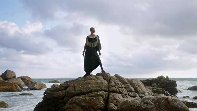 Divine non-binary black person in boutique dress, brass jewelry poses on rocky hill top with dramatic ocean cloudy skyline. Queer lgbtq fashion model in posh outfit fluttering in air. Pantheon concept