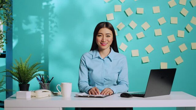 Cheerful business asian employee in bright administration workplace Happy smiling businesswoman working at modern relaxed creative office desk over blue studio background