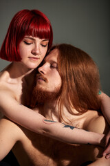 A young attractive woman with bright ruby hair, tattoos and piercings hugs her man with long hair and beard from behind. Half-naked lovers in each other's arms. Close-up studio shot, dark background.