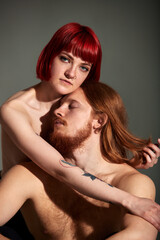 A young attractive woman with bright ruby hair, tattoos and piercings hugs her man with long hair and beard from behind. Half-naked lovers in each other's arms. Close-up studio shot, dark background.