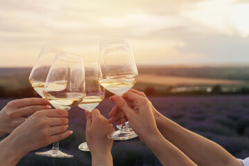 Group of happy girlfriends clinking glasses with wine at picnic on lavender field at sunset.	
