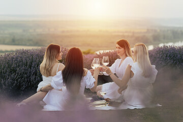 Company of young female friends having fun, drink wine, and enjoy beautiful landscape and sunset at summer picnic.	
