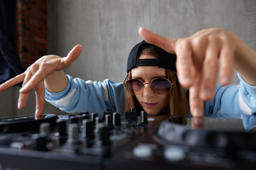 A young pretty long-haired DJ girl in a blue sweater and black baseball cap poses with a black DJ...