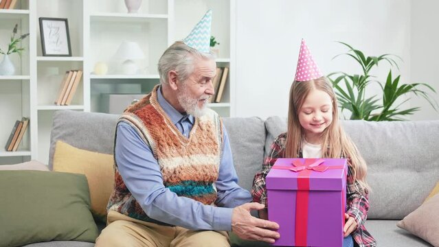 Caring grandfather giving big purple gift box to precious grandchild while celebrating birthday at home. Cheerful young lady rejoicing cool present and clapping hands. Concept of love and surprise.