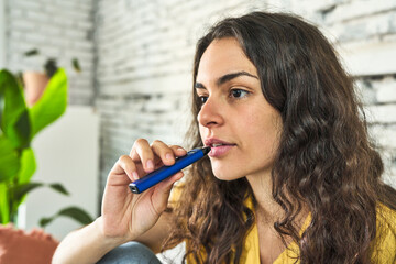 A young Caucasian woman, seated on the sofa, enjoying a vaping device as a healthier alternative to...