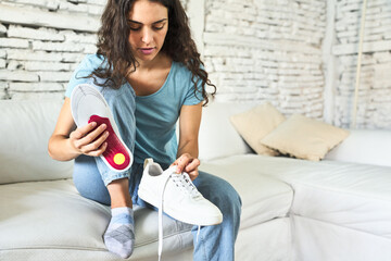 A young Caucasian woman, seated on the sofa, gently placing an orthopedic insole into her shoe, seeking comfort and foot support.