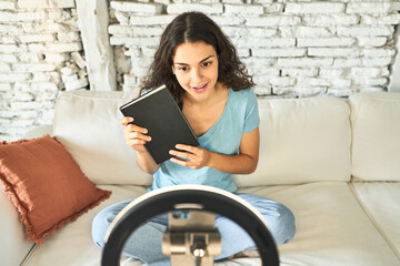 A young Caucasian influencer, sharing book tips and reviews, capturing creative videos on her sofa with a ring light and mobile device.