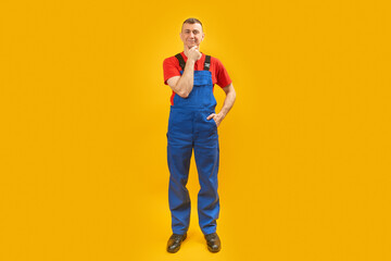 Full-length portrait of builder or worker man in blue jumpsuit put his hand on his chin and thinks. Isolated on yellow background. Copy space.