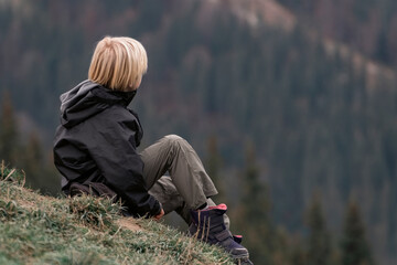Blond boy on hike during break. Child in trekking clothes is relax on the mountainside. Boy sitting on hill