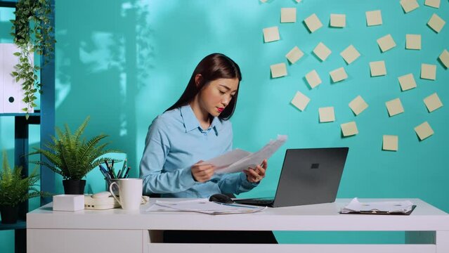 Focused businesswoman executive cross-referencing invoicing administrative data between paperwork and laptop. Professional office clerk working in modern workplace over blue studio background
