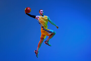 Fototapeta na wymiar Muscular young man in yellow uniform, basketball player during game, jumping with ball against blue studio background in neon light. Concept of professional sport, hobby, healthy lifestyle, action