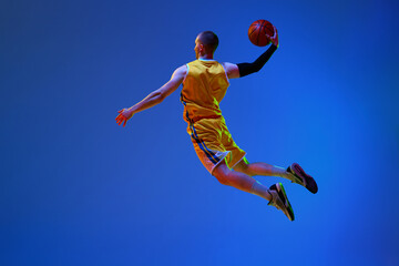 Fototapeta na wymiar Back view image of young male basketball player in yellow uniform training, jumping with ball against blue studio background in neon light. Professional sport, hobby, healthy lifestyle, action concept
