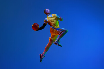 Young man, basketball player in yellow uniform training, throwing ball in a jump against blue studio background in neon light. Concept of professional sport, hobby, healthy lifestyle, action, motion