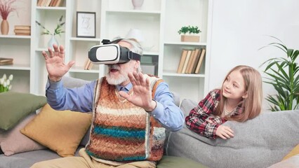Stylish modern grandfather and granddaughter playing together interesting games using virtual reality headset. Excited senior man waving hands frighteningly. Futuristic technology concept. - Powered by Adobe