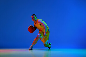 Young man, basketball player in yellow uniform during game, dribbling ball against blue studio...