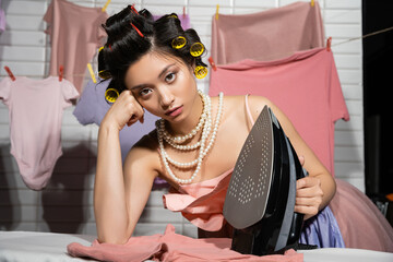 tired asian housewife with hair curlers in pink ruffled top and pearl necklace looking at camera...