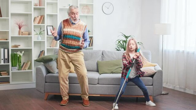 Adorable little girl with cool grandpa dancing while cleaning house. Artistic grandchild in jeans and checkered shirt fooling around with mop. Concept of family and funny leisure activities.