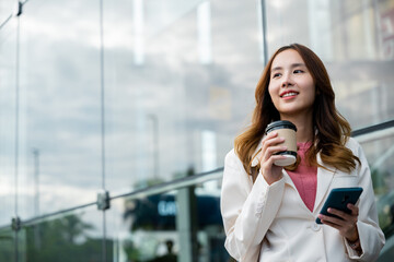 Asian businesswoman with smartphone and cup coffee standing street front building near office,...