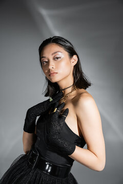portrait of alluring asian young woman with short hair posing in black strapless dress with belt and gloves while looking at camera on grey background, wet hairstyle, golden necklaces
