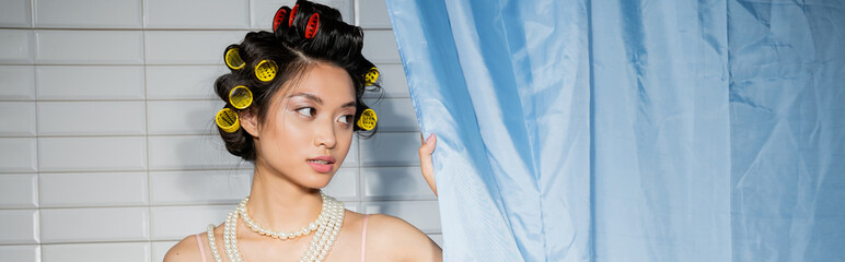 brunette and alluring asian woman with hair curlers standing in pearl necklace near blue bathroom...