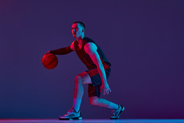 Young muscular man, basketball player in motion, dribbling ball against purple studio background in...