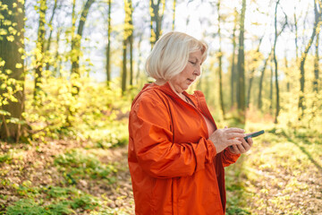 Middle aged woman holding mobile phone in hands outdoors. Beautiful mature woman 50s in green sporty wear jogging in park and smiling, looking at her mobile phone app,