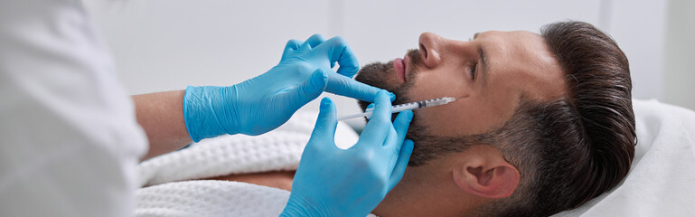 Mature man patient lies on couch undergoing crows feet removal procedure in clinic