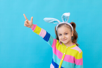 portrait of a little girl with bunny ears in a striped jacket smiles and points her fingers up. The concept of the Easter holiday. Blue background, photo studio, place for text