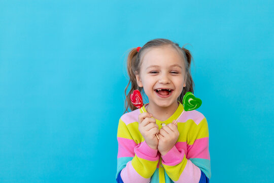 portrait of a joyful little girl with lollipops in a striped jacket, a large candy on a stick. The concept of sweets and confectionery. Blue background, photo studio, place for text