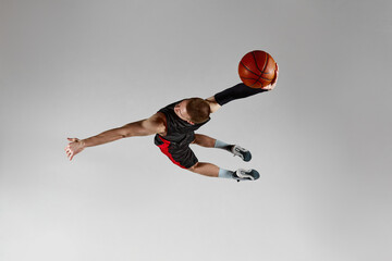 Winning goal. Sportive young man, basketball player in motion, jumping with ball against grey...