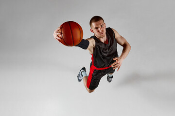 Concentrated sportsman, male basketball player in motion, jumping with ball against grey studio...