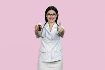 Asian female doctor is giving medicine bottle and her thumb up. Isolated on pink background.