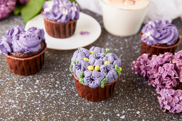 Fototapeta na wymiar Floral purple cupcakes using trend Dreamy Escapism. Aesthetics desserts, coffee cup and lilac flowers background. Catering food