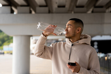 Fototapeta na wymiar Tired Asian man with smartphone in his hands is drinking water after training outdoors