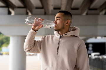 A young guy drinks water from a bottle to quench his thirst after a long run. A young Asian man...